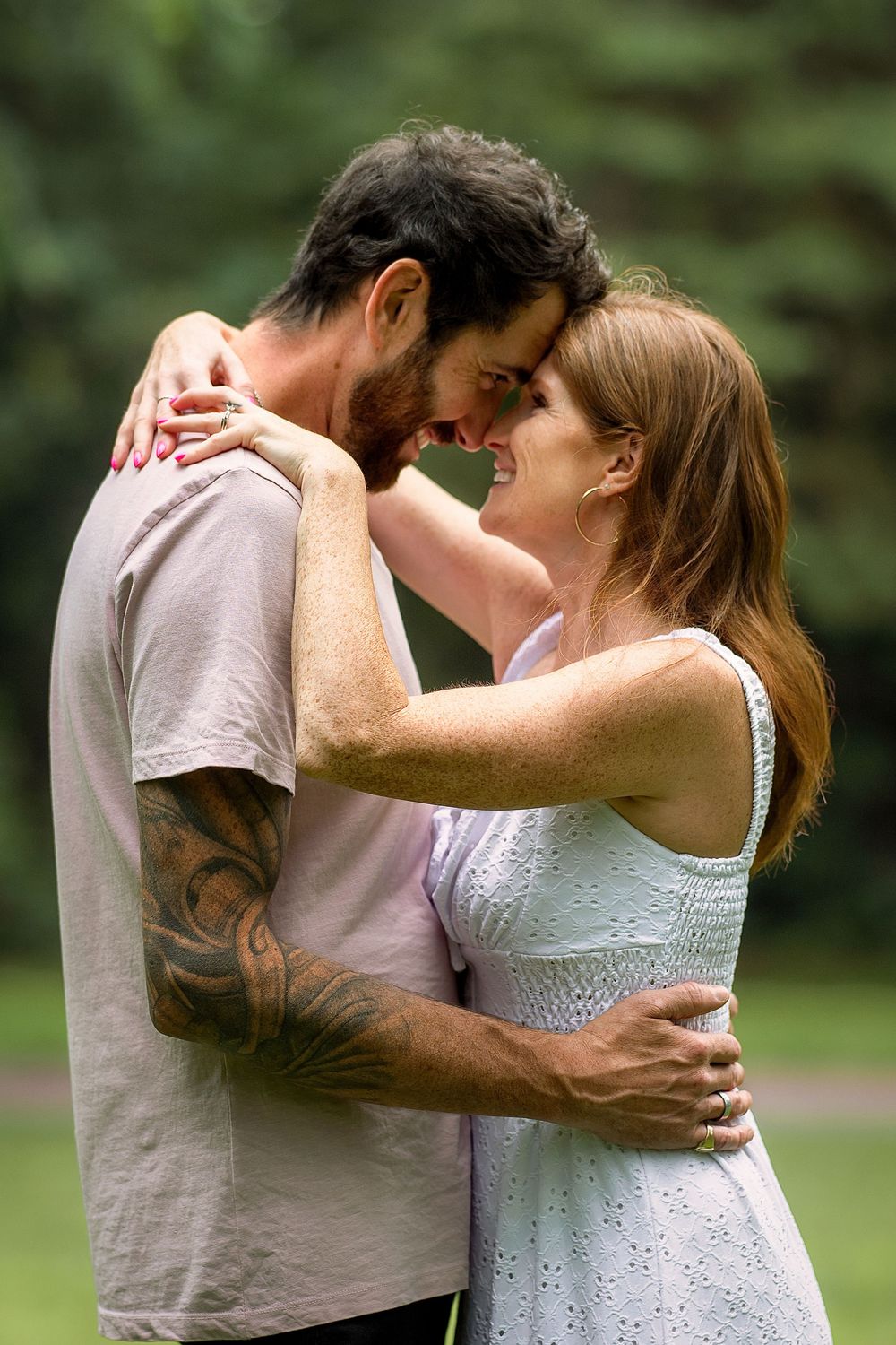 A couple embraces forehead-to-forehead, smiling in a lush green park, man with tattoos in gray tee, woman in white dress.