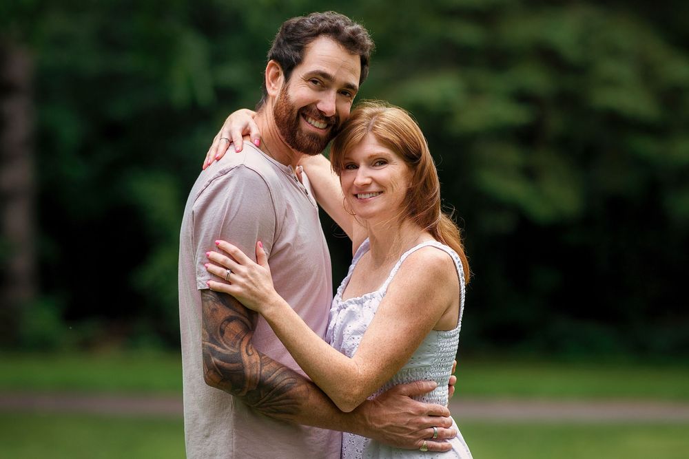 A smiling couple embraces outdoors in a lush green park, the man with tattoos and the woman in a sleeveless top.