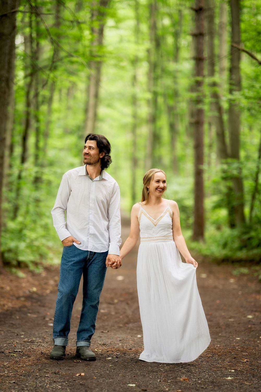 A couple holds hands on a forest path, the woman in a white dress and the man in jeans and a white shirt.