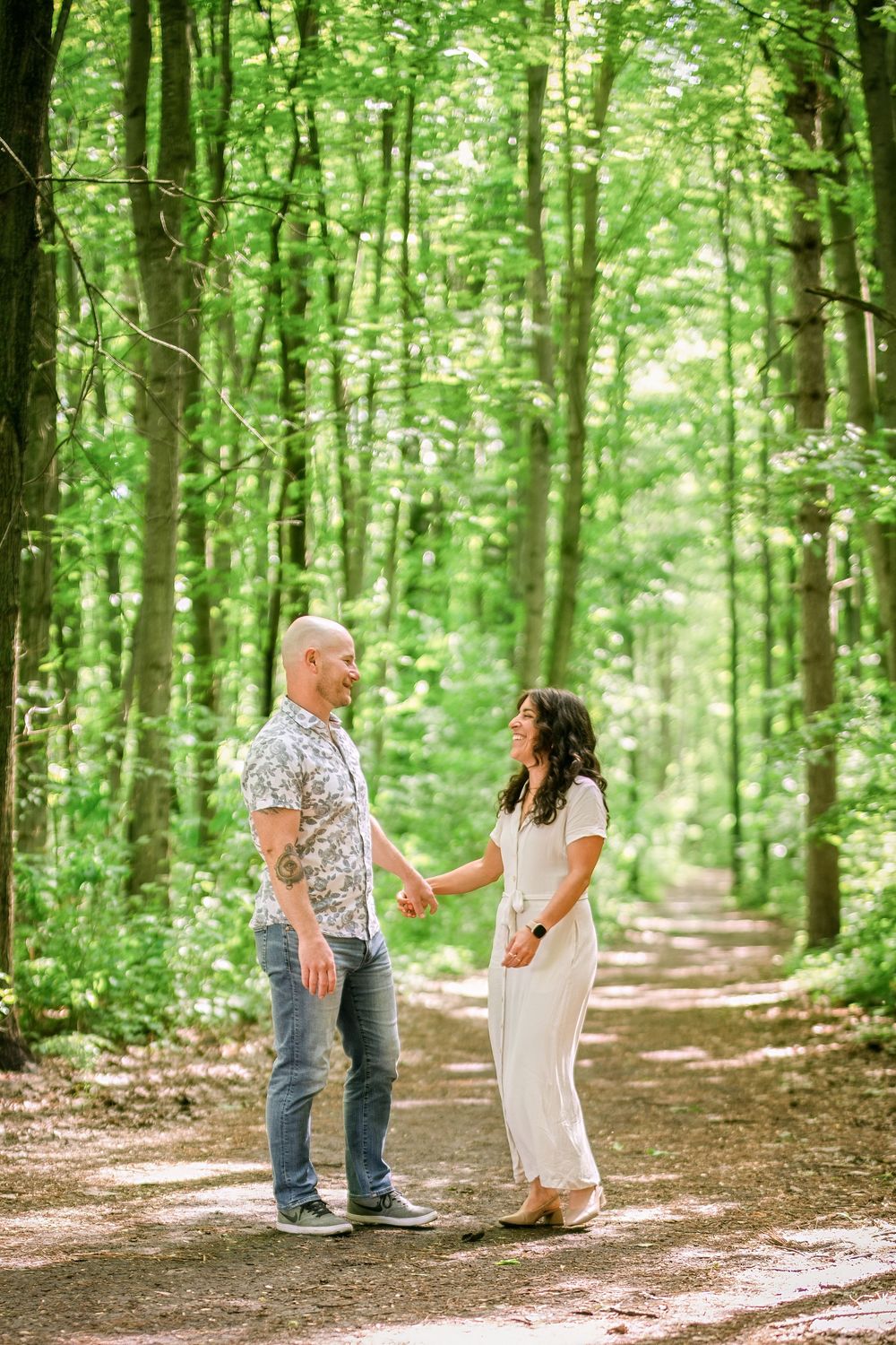 Couple holding hands on a sunlit forest path surrounded by lush green trees.