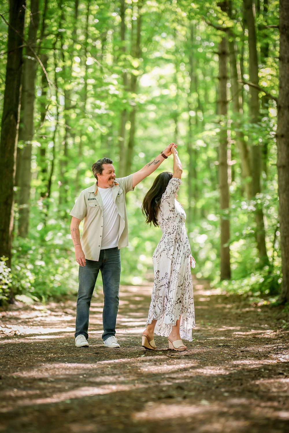 A couple dances joyfully on a sunlit forest path, the woman in a floral dress spinning under the man's raised hand.