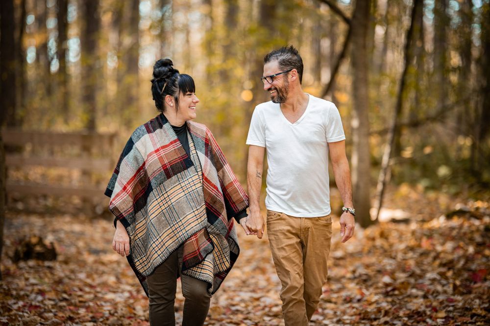 Couple walking hand in hand on a forest trail in autumn, surrounded by golden fallen leaves and bare trees.