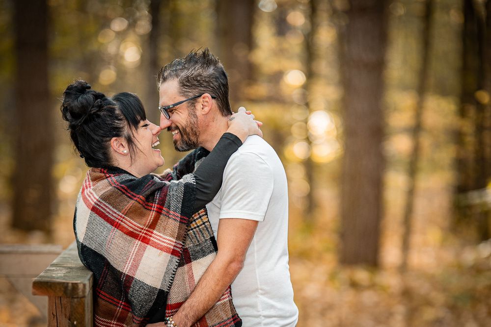 Couple embracing and smiling in autumn forest, woman wrapped in plaid blanket, man in white t-shirt.