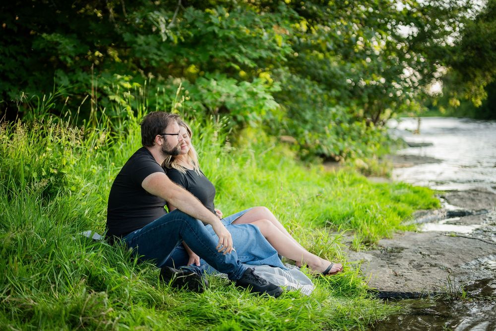 Couple sitting together on green grass beside a peaceful stream surrounded by lush trees.