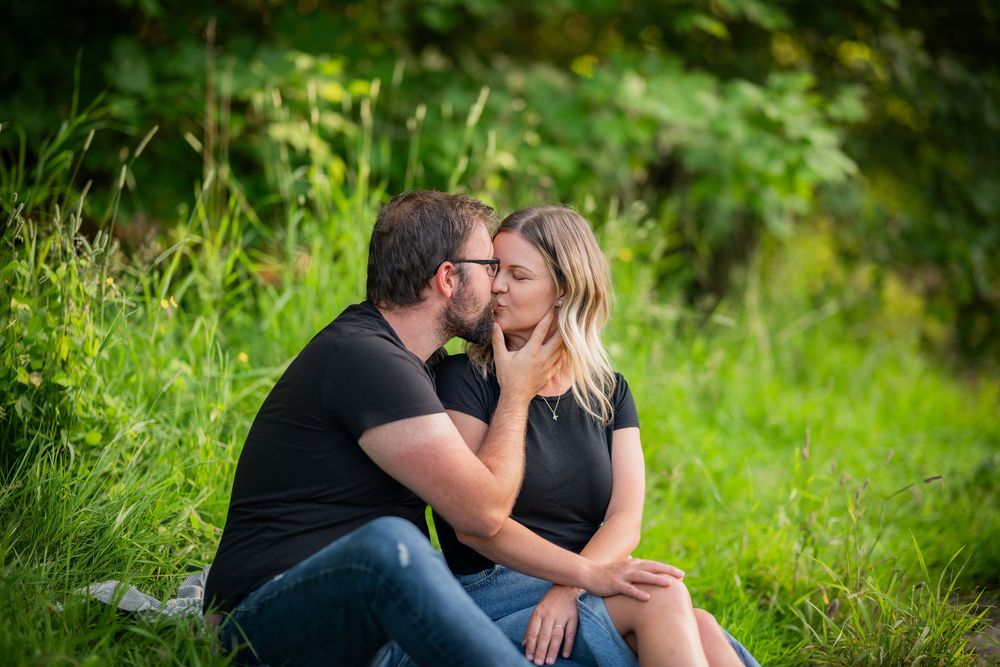 A couple shares a romantic kiss while sitting on green grass surrounded by lush foliage outdoors.