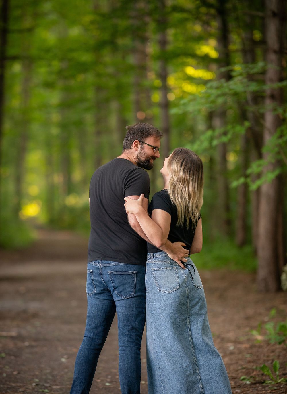 A couple shares a romantic moment on a forest path, embracing and smiling at each other surrounded by lush green trees.