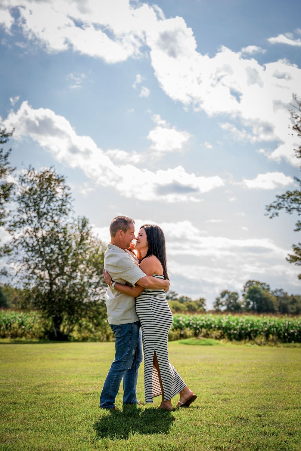 Couple embracing and almost kissing on a green lawn with trees and a blue cloudy sky backdrop.