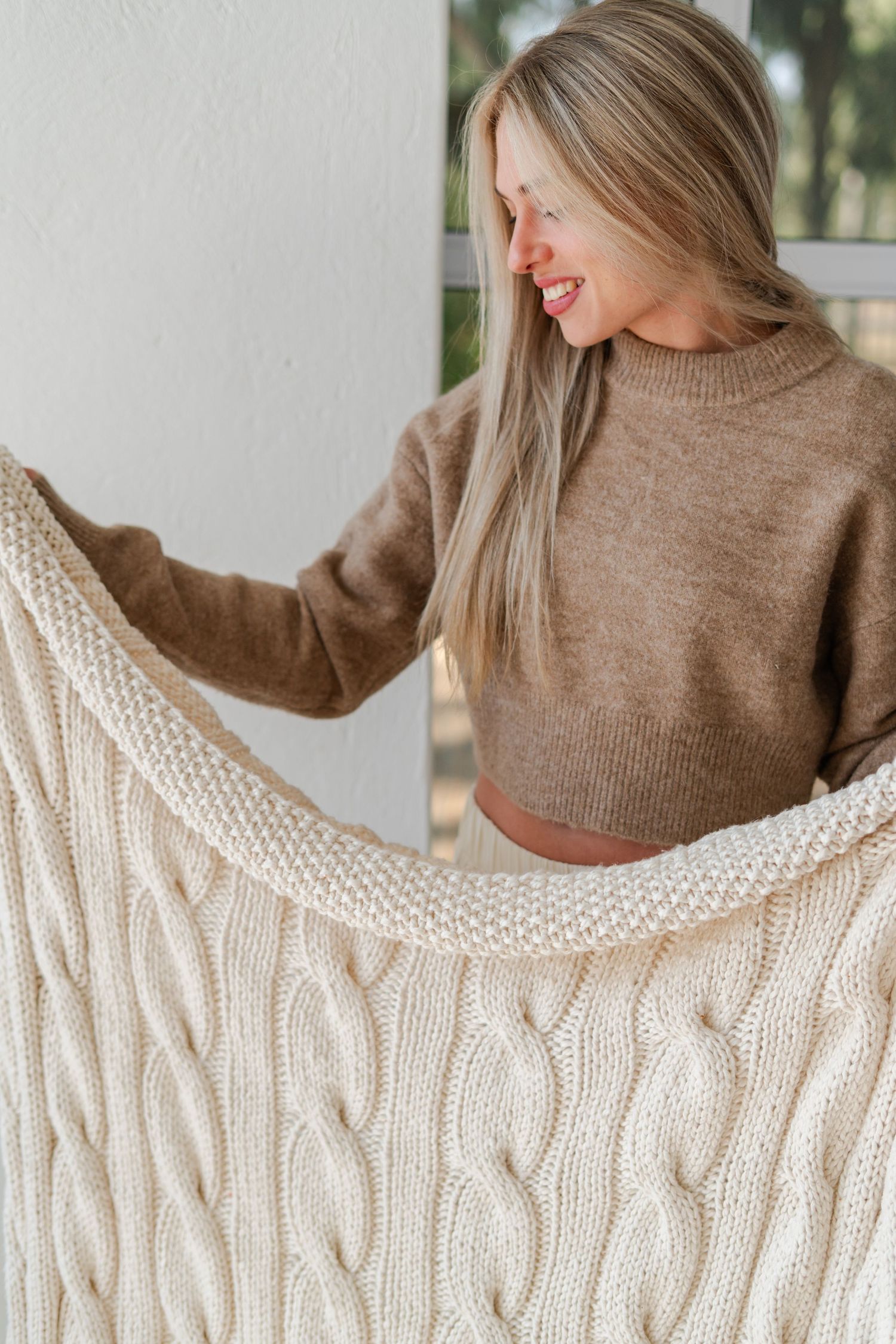 Smiling blonde woman in brown cropped sweater holding a cream cable-knit LYBL throw during their brand product photoshoot.