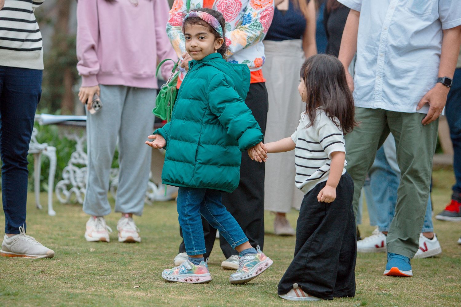 Two young children in colorful outfits hold hands on green grass at an outdoor event with adults nearby.