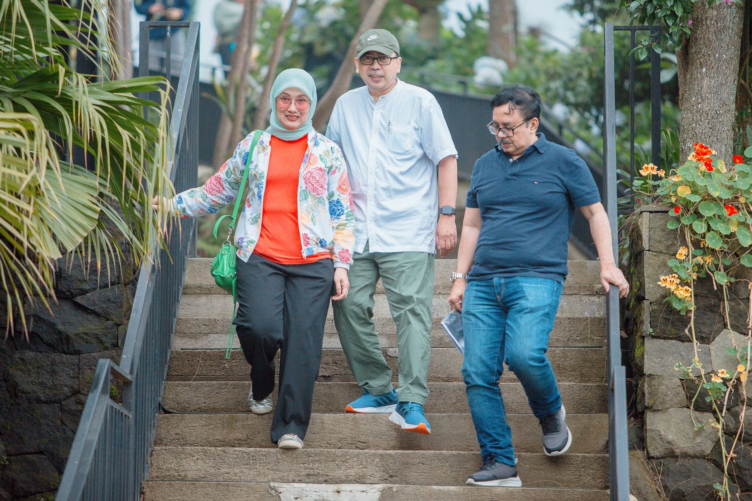 Three people walking down outdoor stone stairs surrounded by tropical plants and flowers.