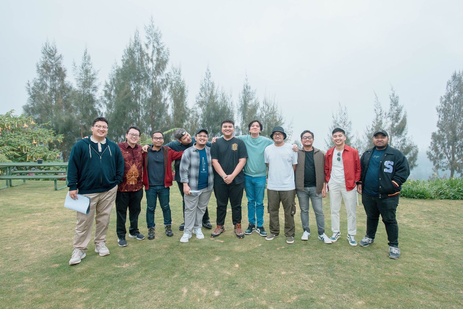 Group of eleven men posing together on green lawn with tall pine trees and misty sky backdrop.