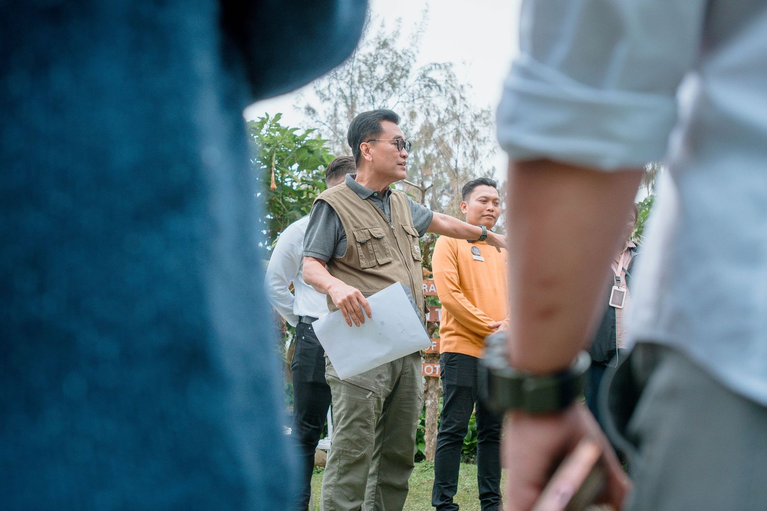 A man in a khaki vest holds documents while speaking to a group outdoors during a field discussion.