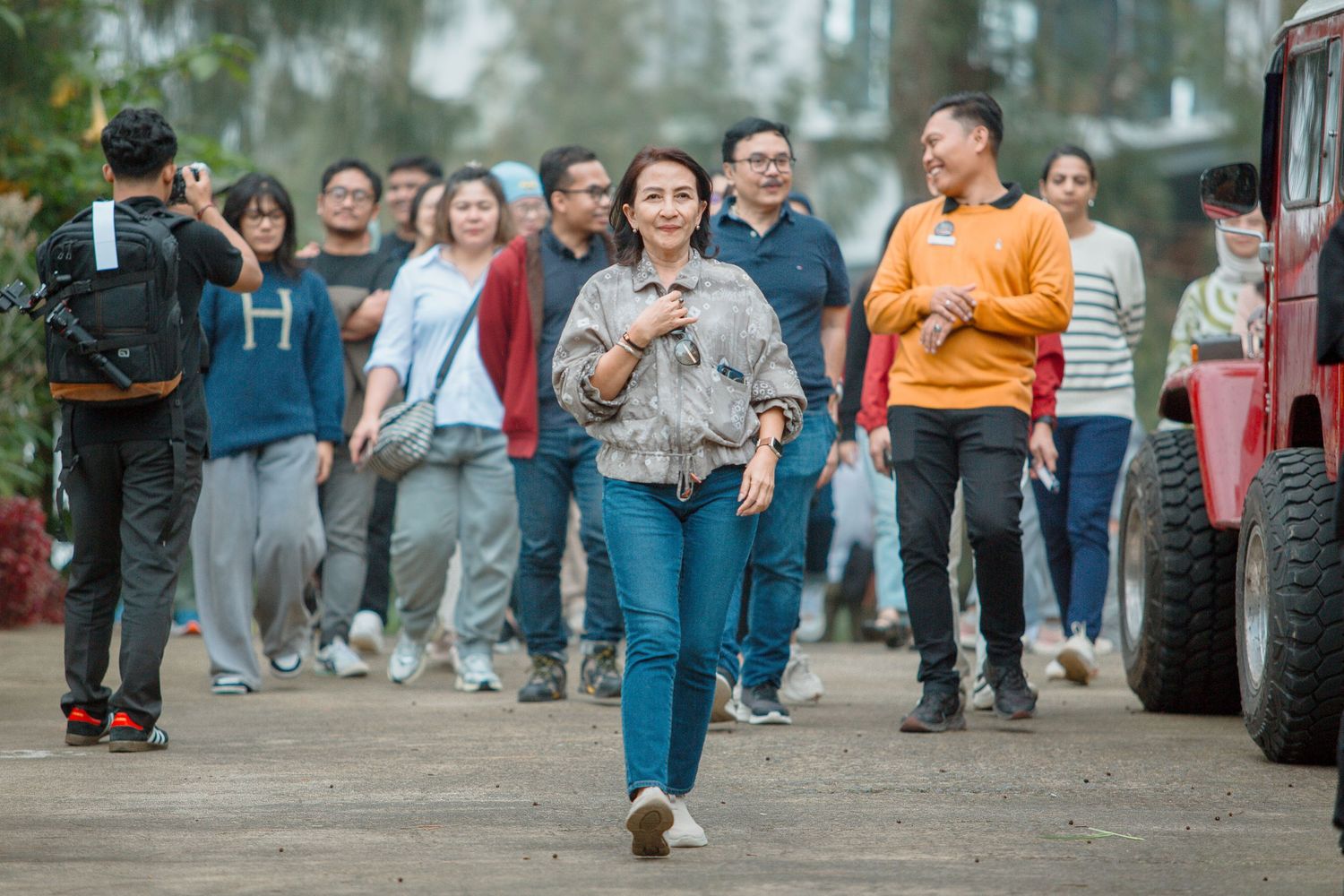 Woman in denim jacket walks confidently ahead of a crowd outdoors near a red vehicle.