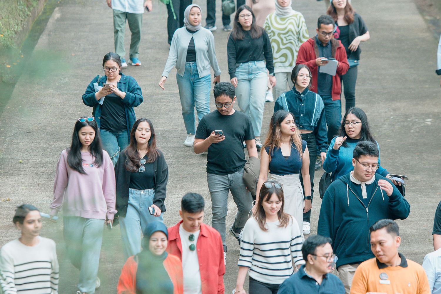 A diverse crowd of young people walking outdoors on a path, some using smartphones, in casual attire.