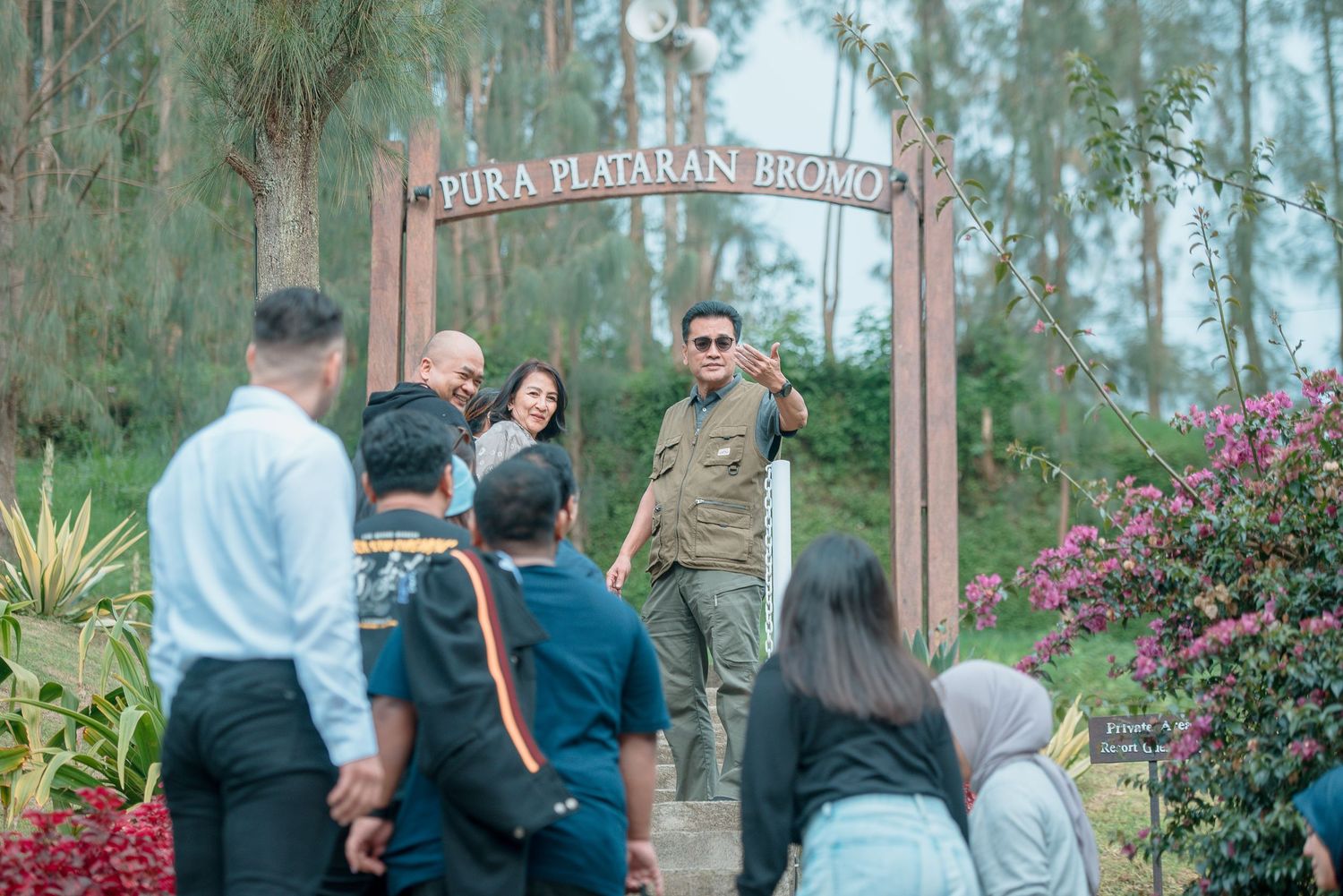 Tourists gather at Pura Plataran Bromo entrance gate surrounded by lush greenery and colorful flowers in Indonesia.