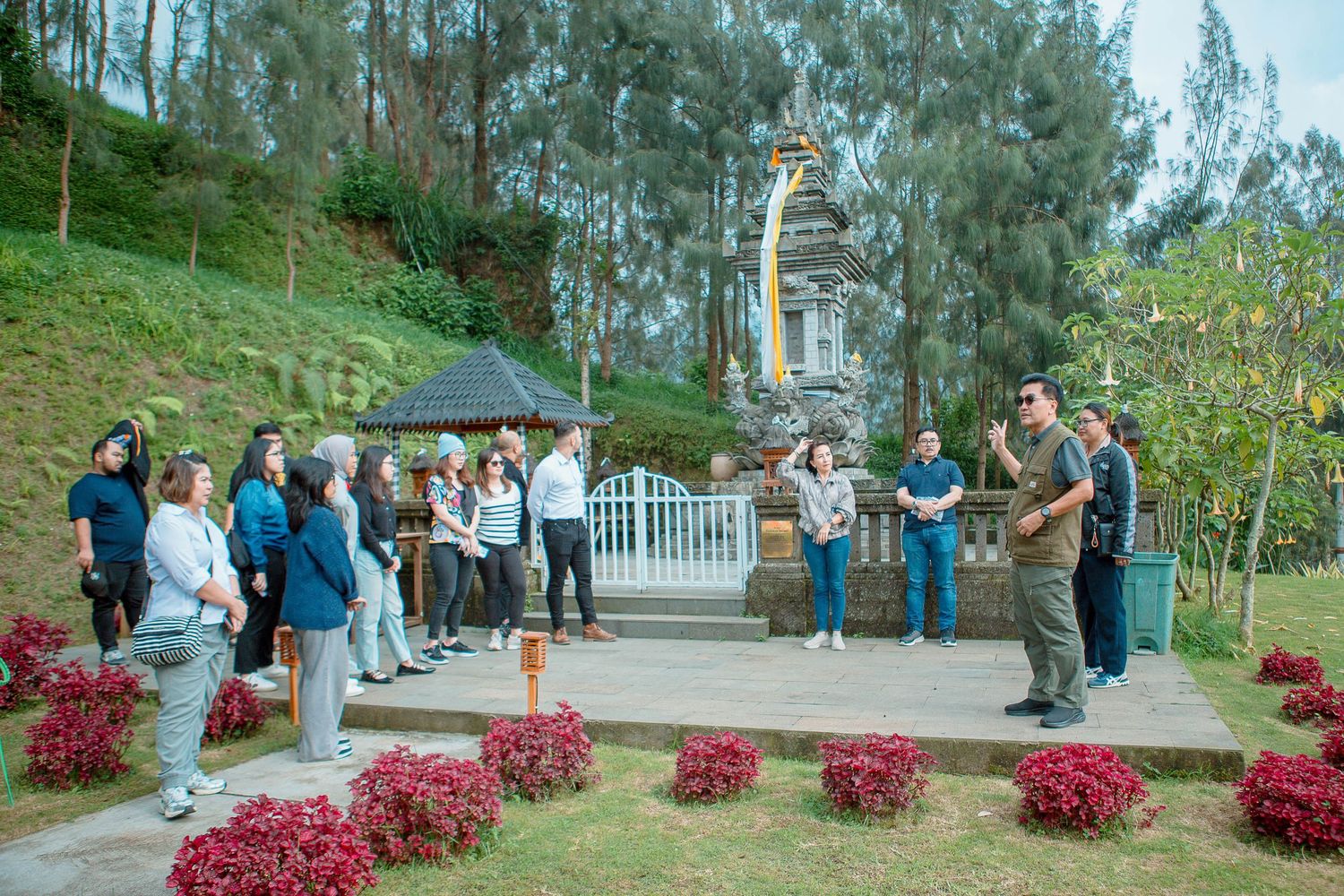 A group of people gathered around a monument in a lush garden with trees and red plants nearby.