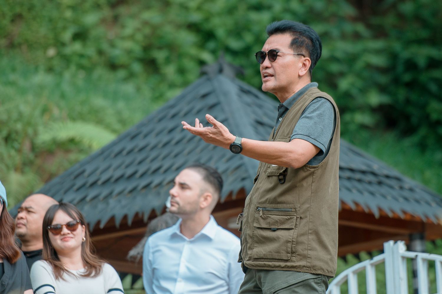 Man in olive vest speaking gesturing outdoors with group of people seated behind him near a wooden gazebo.