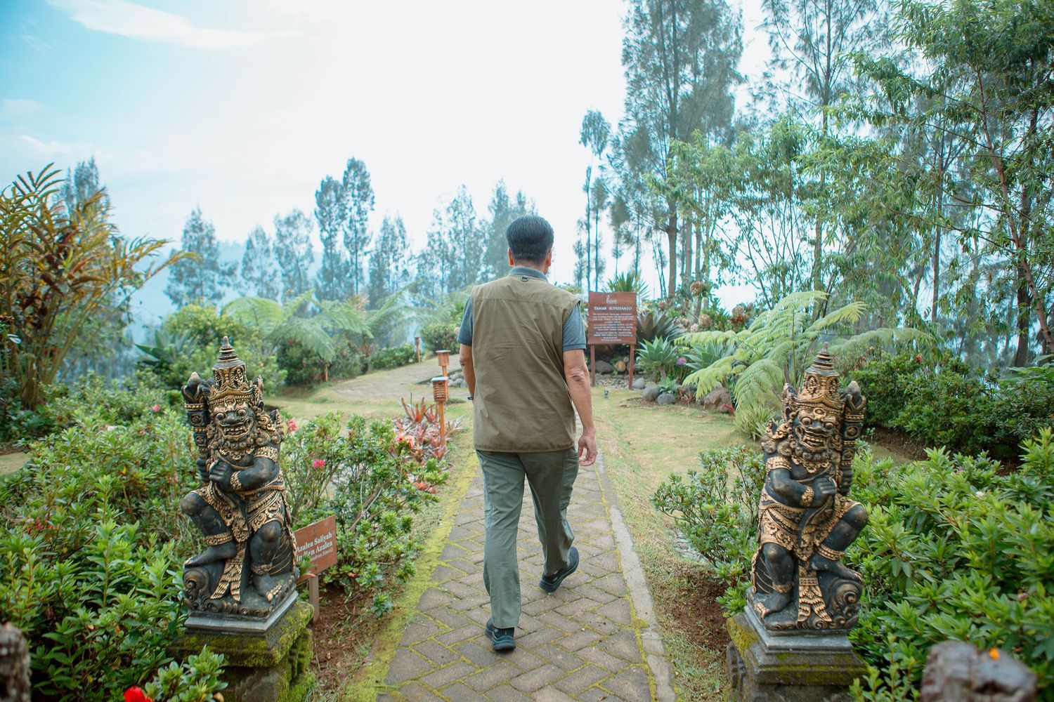 Man walking through a lush garden pathway flanked by ornate stone statues and tropical greenery.