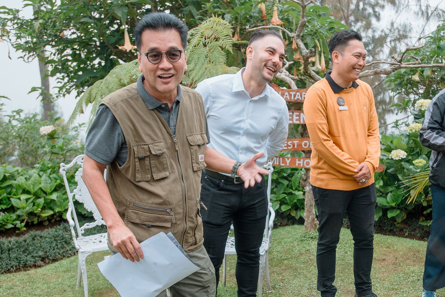 Three men laughing and socializing outdoors at a garden event, dressed in casual and semi-formal attire.