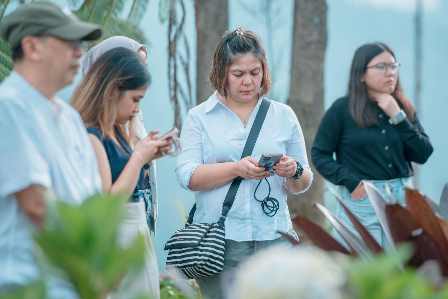 Group of people standing outdoors looking at their smartphones near tropical plants with ocean view.