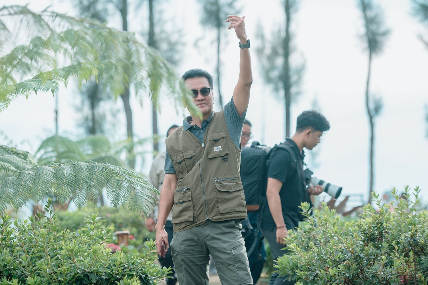 Man in olive vest raises hand outdoors, surrounded by lush tropical greenery and companions with cameras.