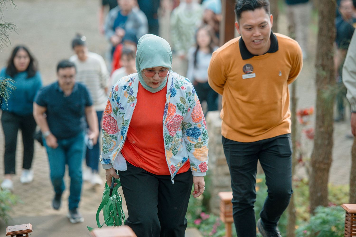A woman in a floral jacket and hijab walks with a man in an orange shirt through a crowded outdoor park.