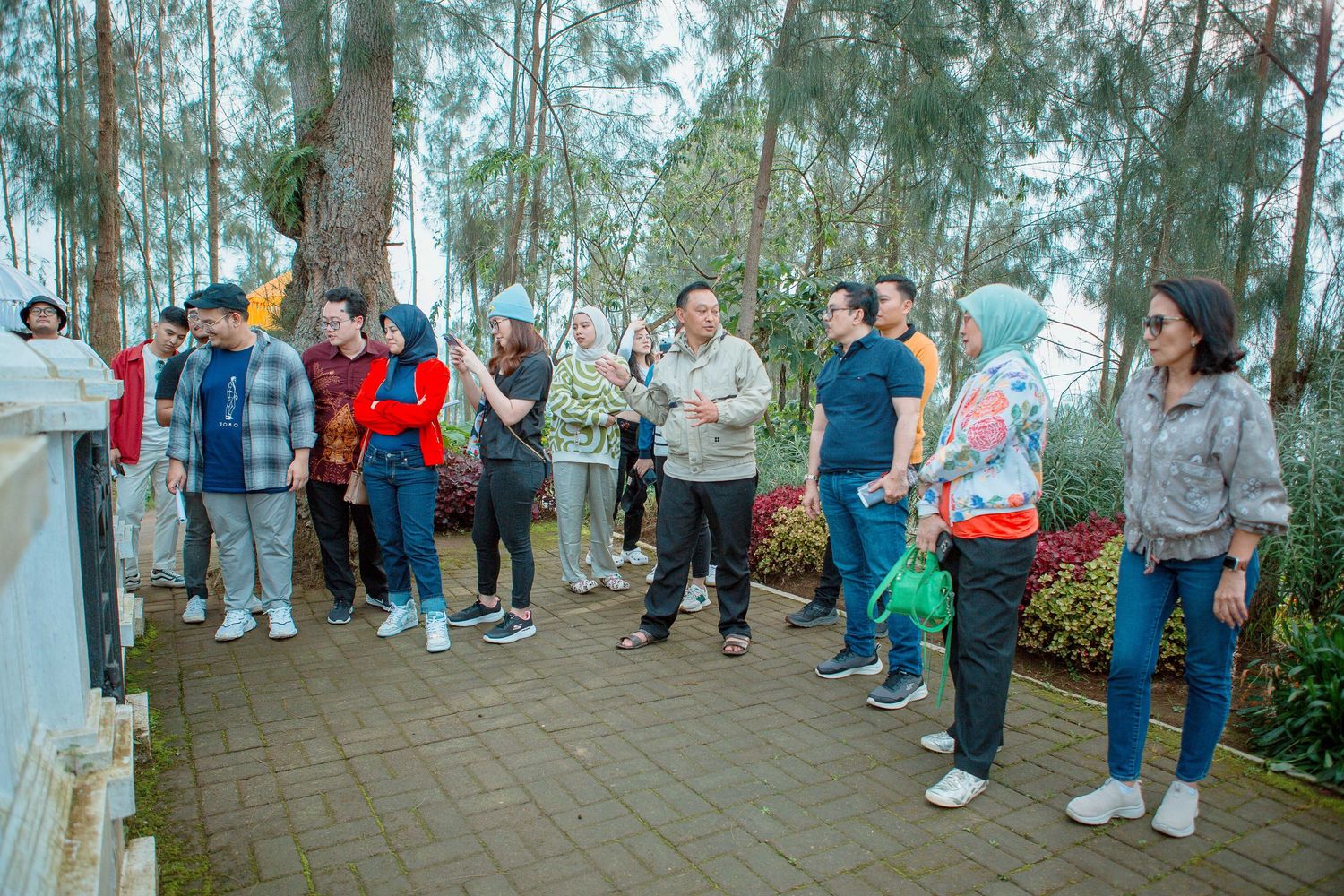 Group of tourists standing on a paved path surrounded by pine trees and colorful flowers at an outdoor park.