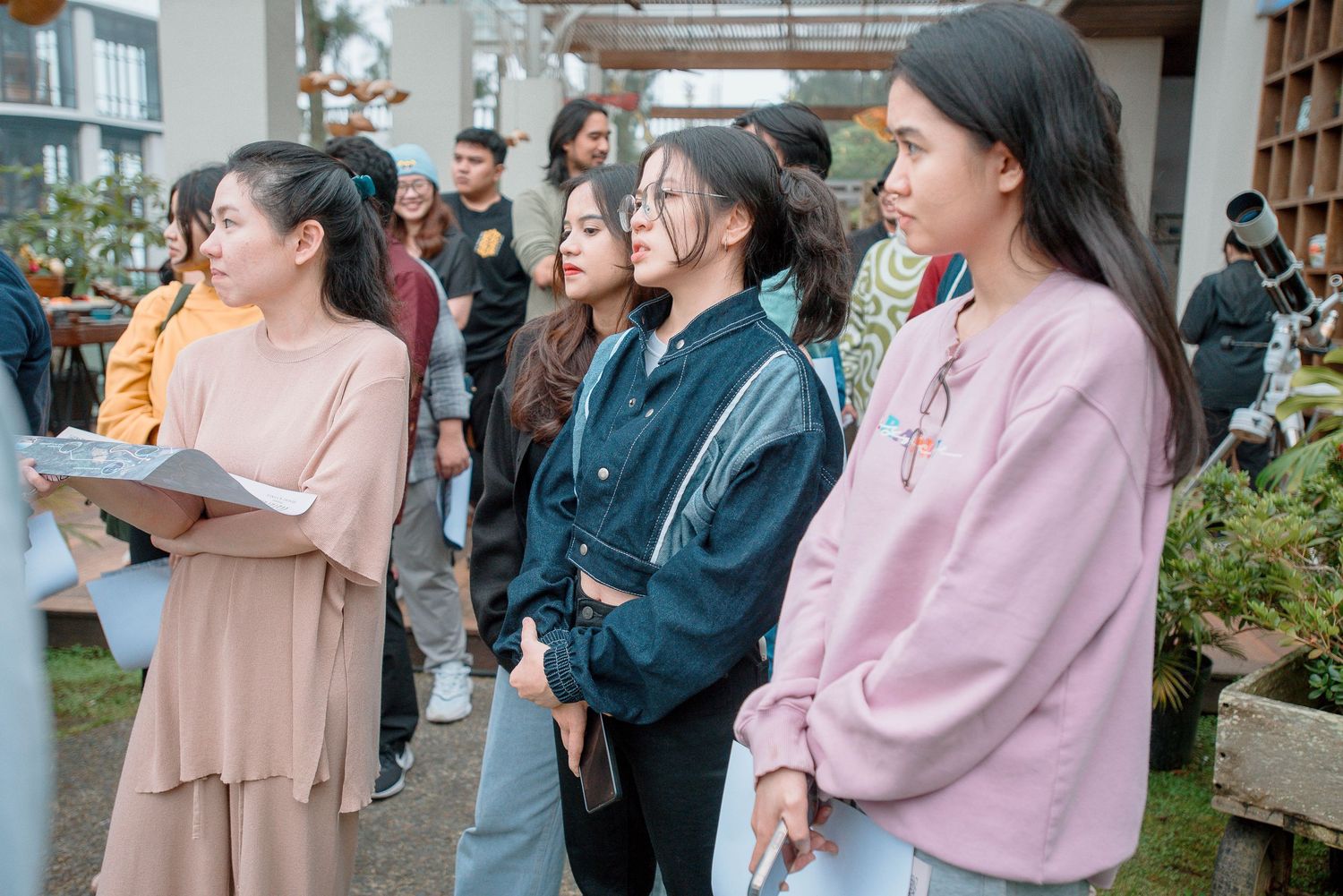 A group of young Asian women standing outdoors at an event, listening attentively in casual colorful clothing.