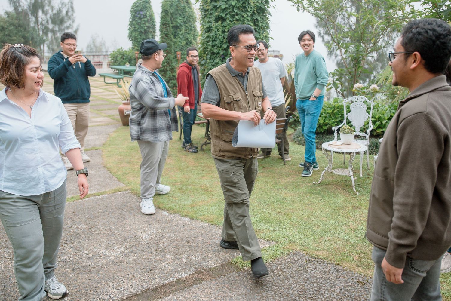 Group of people gathered outdoors on a green lawn near a garden path, engaging in conversation on an overcast day.