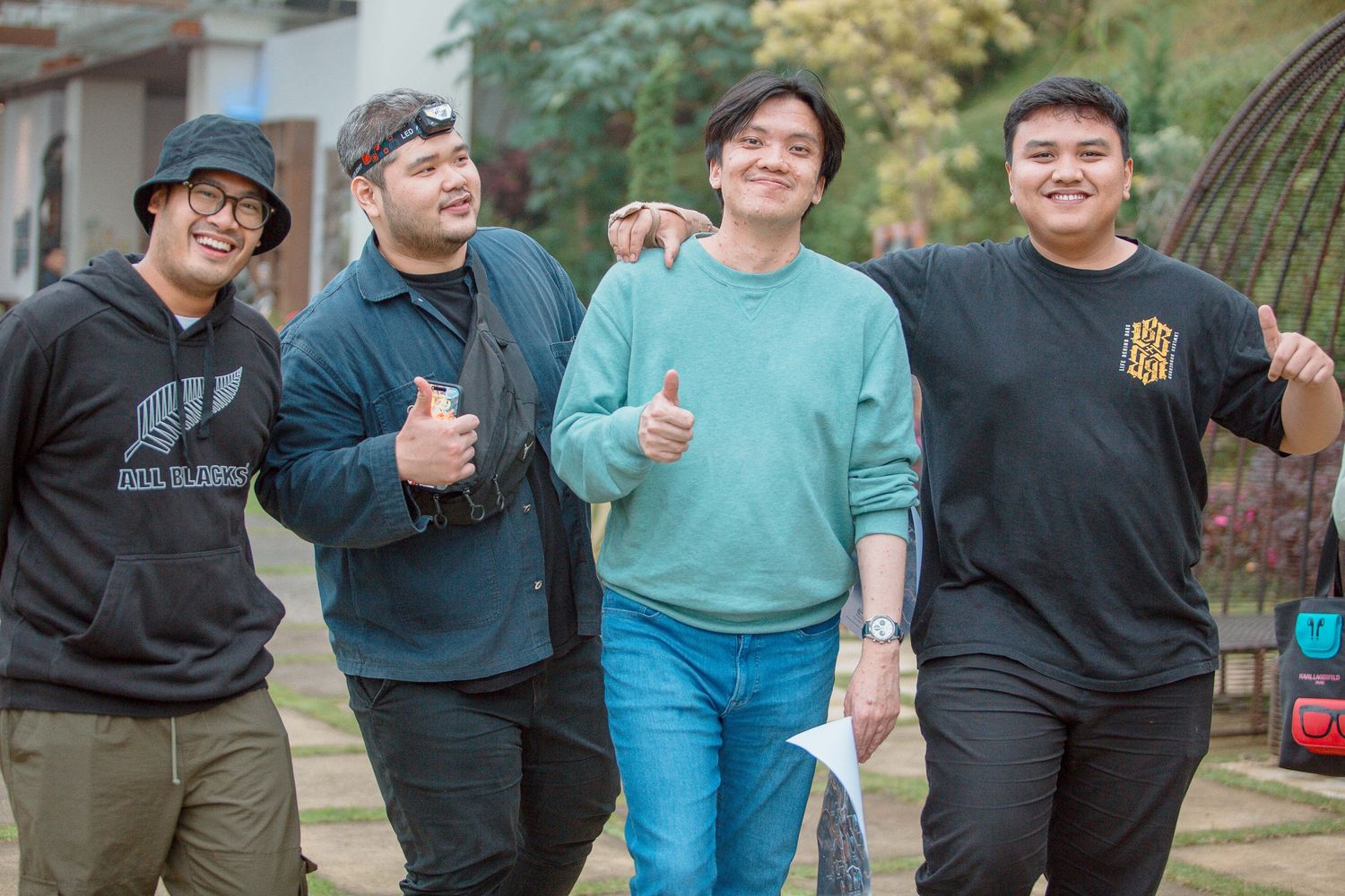 Four young men smiling and giving thumbs up outdoors, wearing casual sweatshirts in a garden setting.