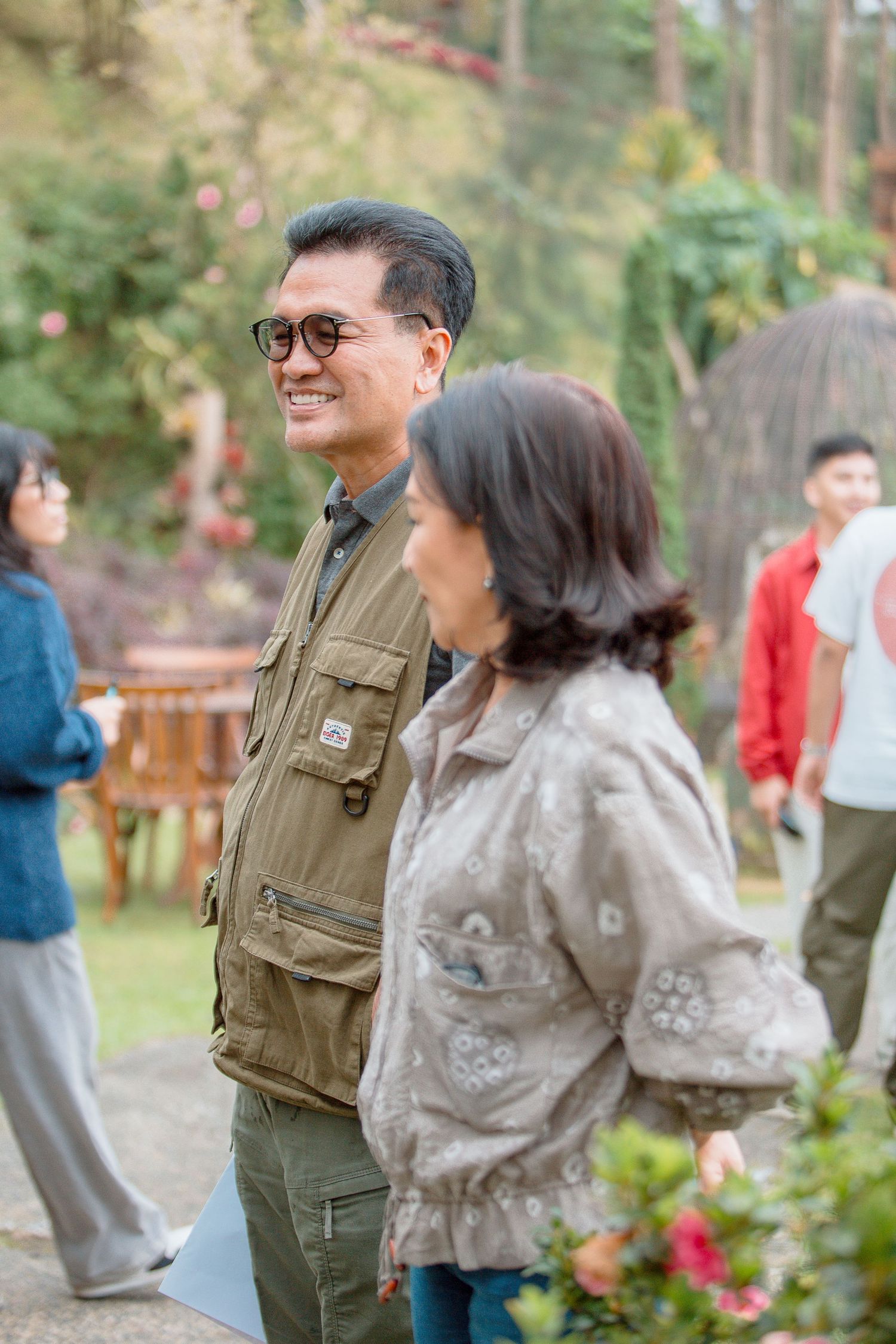 A smiling man in sunglasses and olive vest stands outdoors with a woman in a floral jacket at a garden gathering.