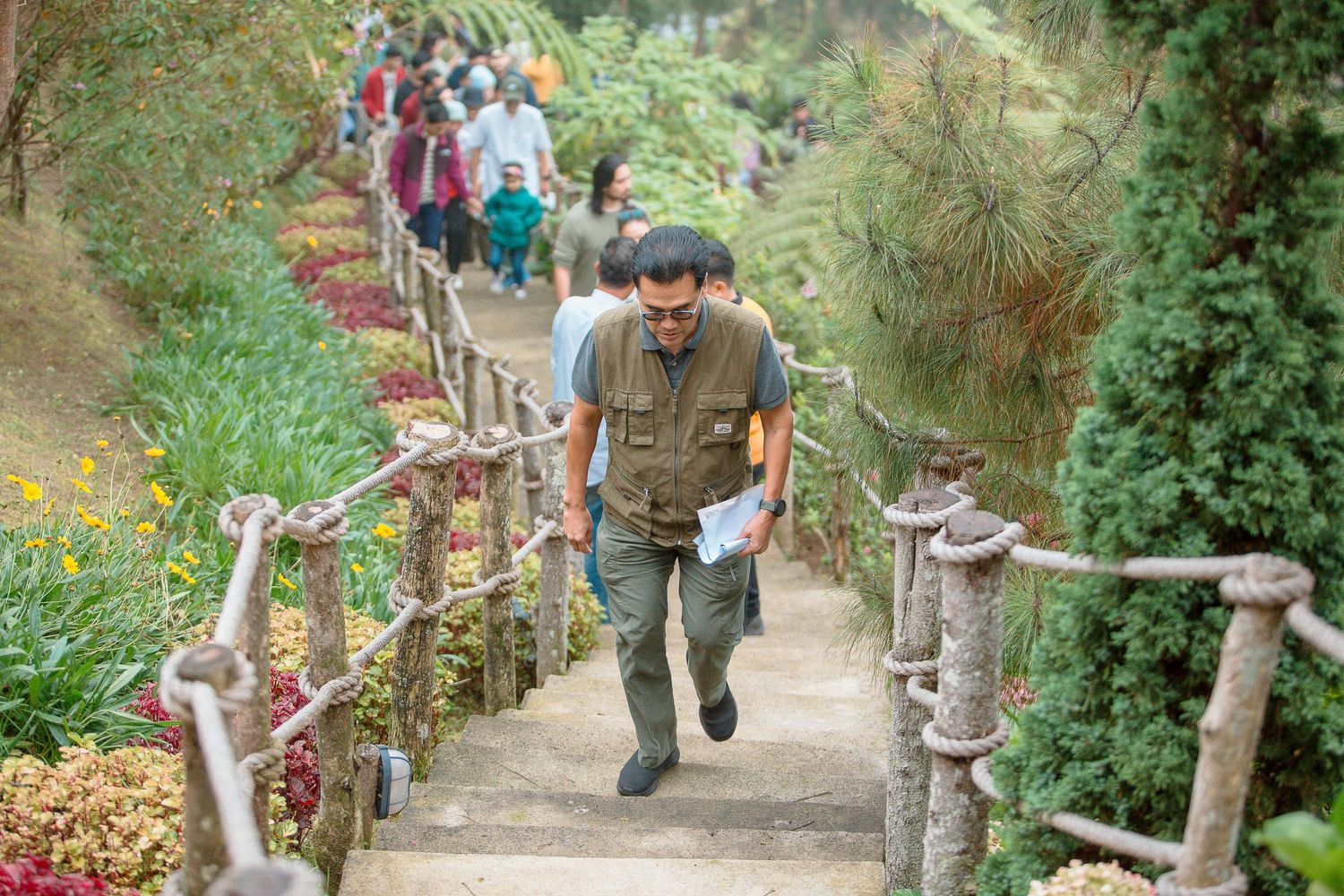 Man in vest climbing stone steps with rustic wooden railings in a colorful hillside garden.