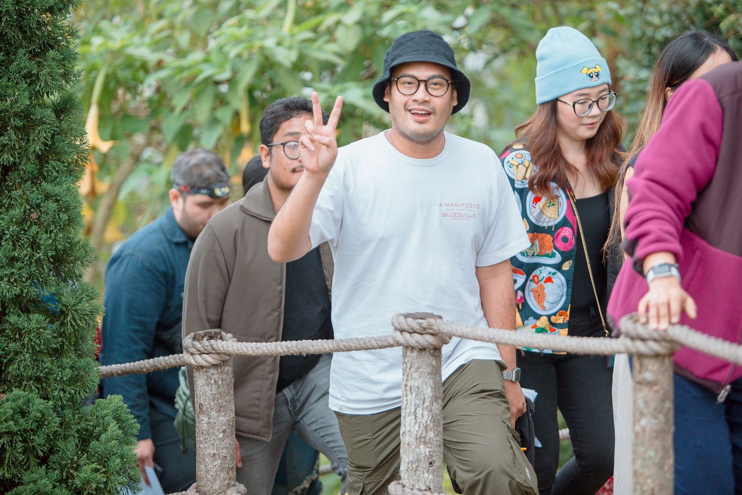 Smiling man flashing peace sign on rope bridge with friends in casual outdoor setting.