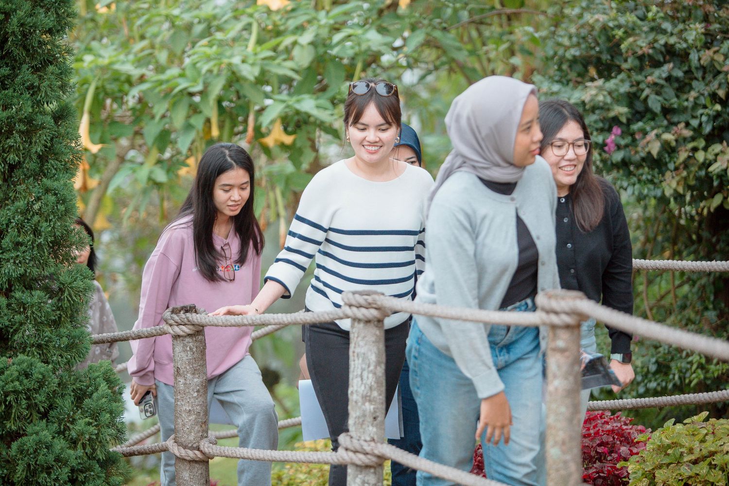 Four young women walking together on a wooden bridge path surrounded by lush green trees in a park.