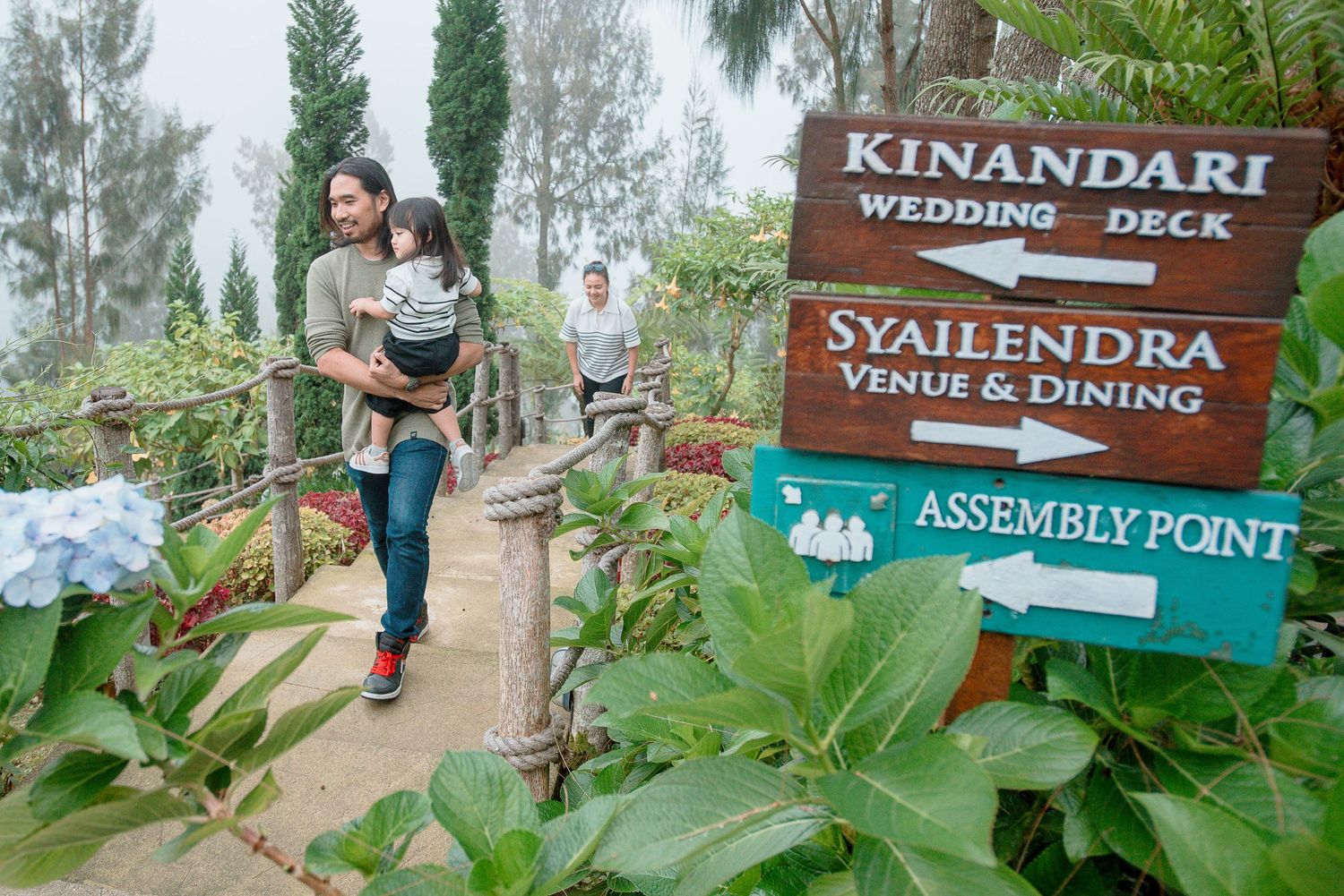 Visitors walk past directional signs for Kinandari Wedding Deck and Syailendra Venue and Dining at a lush garden.