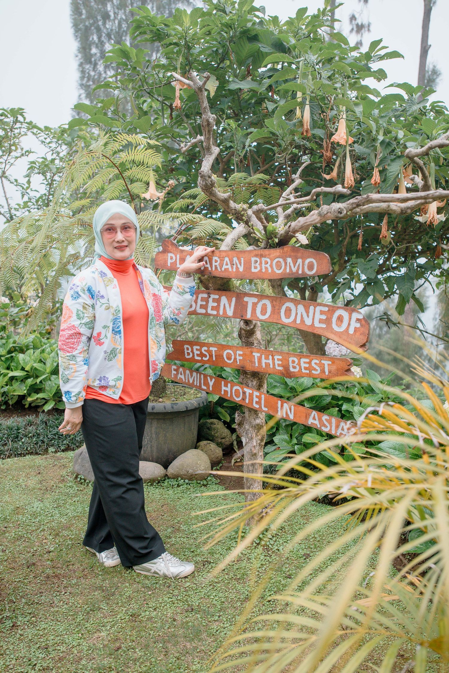 Woman in hijab posing by Plataran Bromo Best Family Hotel in Asia wooden sign surrounded by lush greenery.
