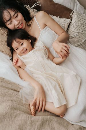 A smiling mother and young daughter in white dresses lying together on a bed, sharing a joyful moment.