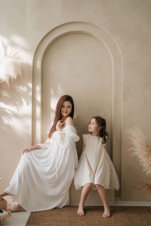 A woman and young girl in matching white dresses pose together against a beige arched wall with pampas grass decor.
