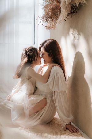 A mother and young daughter in white dresses sit together by a sunlit window, sharing a tender nose-to-nose moment.