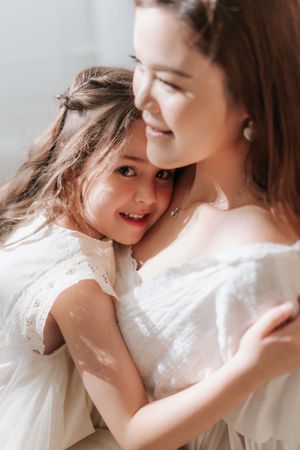 A smiling mother in white dress tenderly embraces her happy young daughter, both sharing a warm and joyful moment.