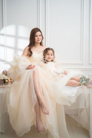 A mother and daughter in matching cream tulle gowns sit together in a bright white room, smiling warmly at the camera.