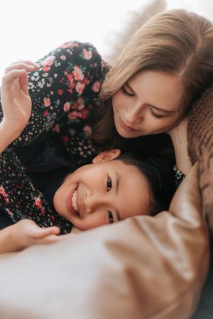 A smiling young girl lying on a couch while a blonde woman lovingly leans over her in matching floral outfits.