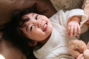 A happy toddler in a white knit sweater smiles while holding a teddy bear, lying back in a cozy setting.