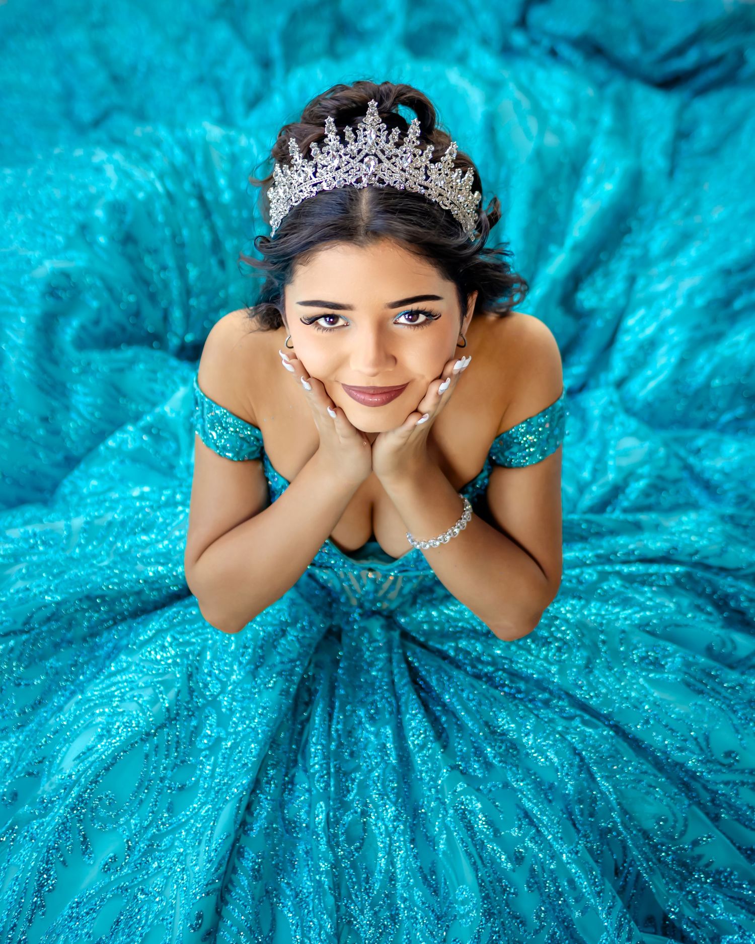 Young woman in a sparkling teal quinceañera gown and crystal tiara poses elegantly with hands under chin.