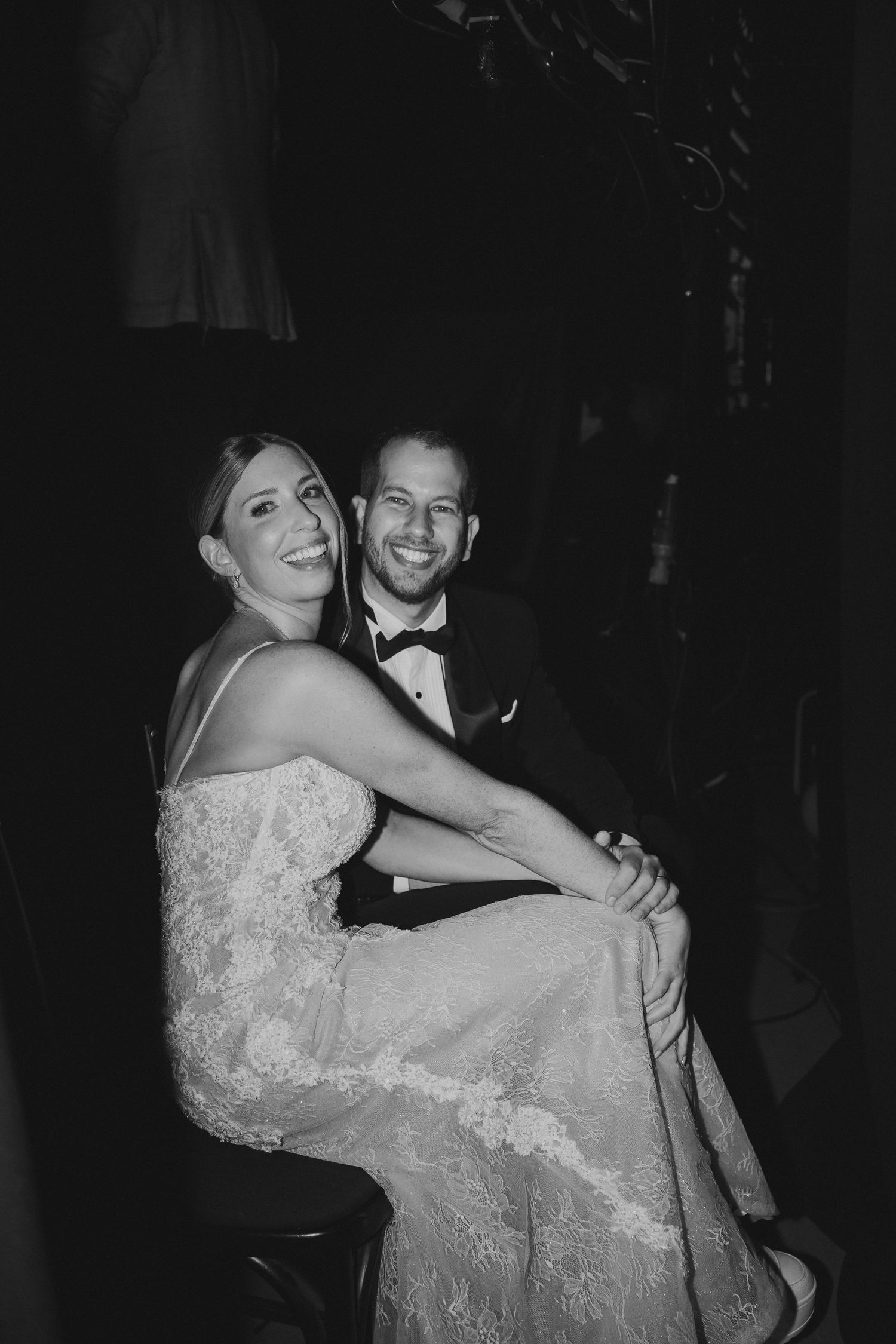 Black and white photo of a smiling bride in lace gown and groom in tuxedo sitting together at their wedding reception.
