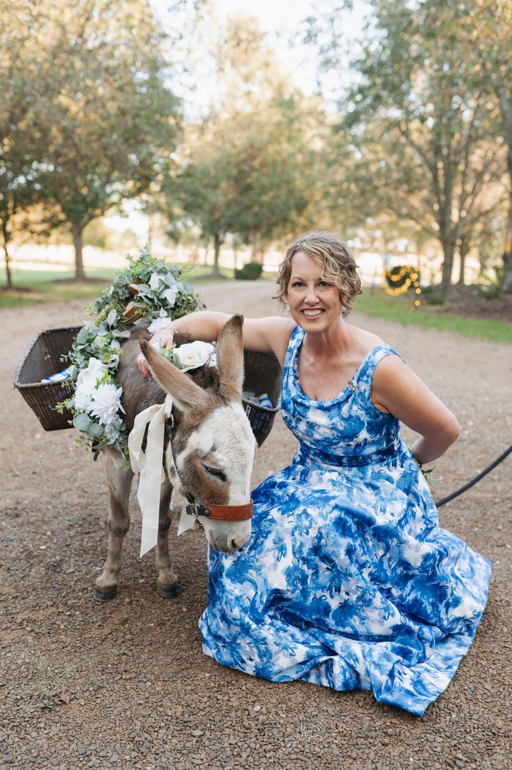 Woman in blue floral dress poses with a decorated donkey at an outdoor wedding celebration.