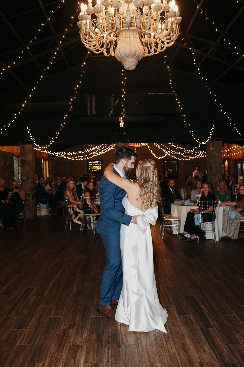 Bride and groom share first dance under chandelier and fairy lights at elegant wedding reception.