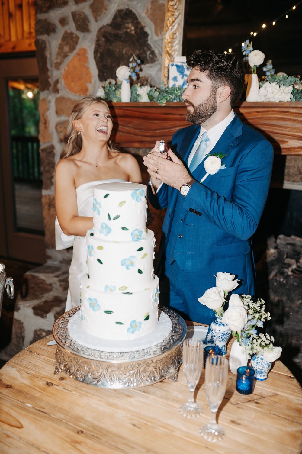 Bride and groom cutting a three-tier white wedding cake with blue floral details at their reception.