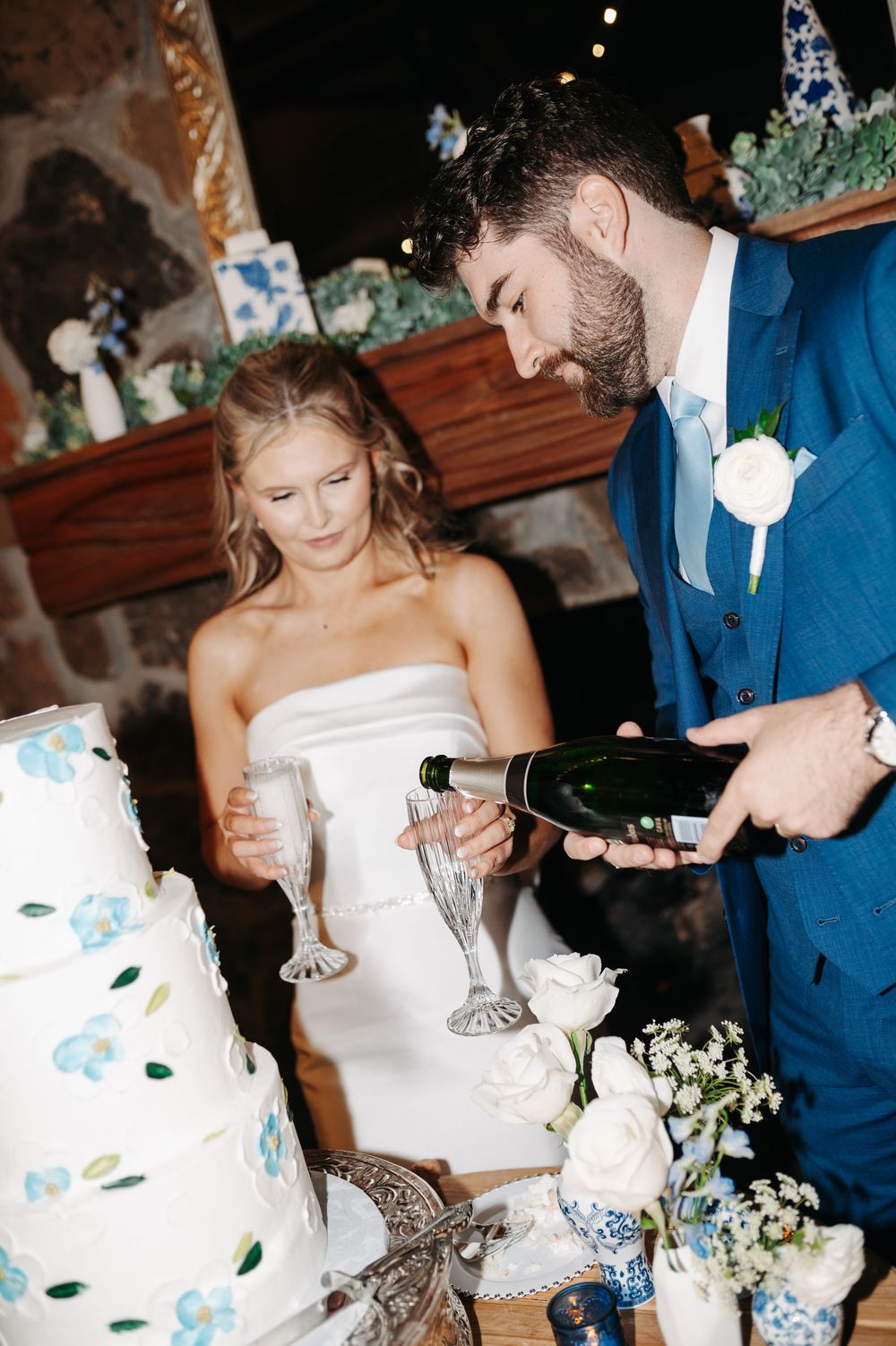 Bride and groom pouring champagne into crystal flutes beside a white wedding cake with blue floral details.