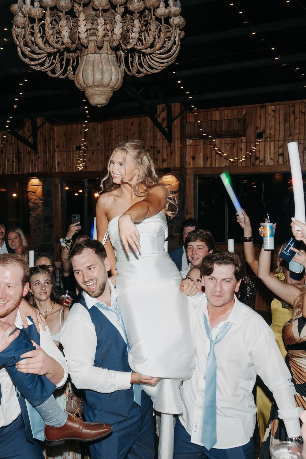 Bride lifted by groomsmen during lively wedding reception with glow sticks and chandelier backdrop.